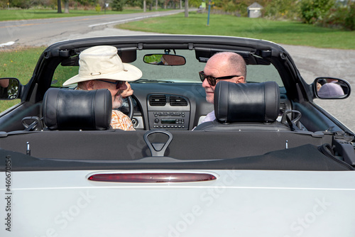 A mature senior gay married couple sit in their white convertible with the top down and look back over their shoulders.  
