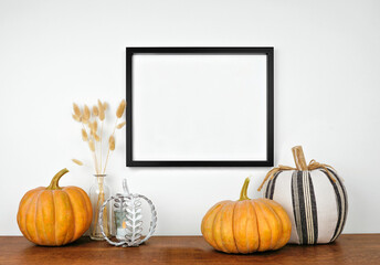 Mock up black frame hanging from a white wall with pumpkins and fall decor on a wood shelf. Autumn concept. Copy space.