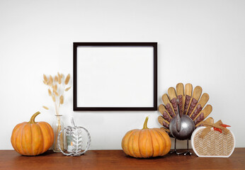 Mock up black frame hanging from a white wall with thanksgiving turkey, pumpkin and autumn decor on a wood shelf. Fall concept. Copy space.