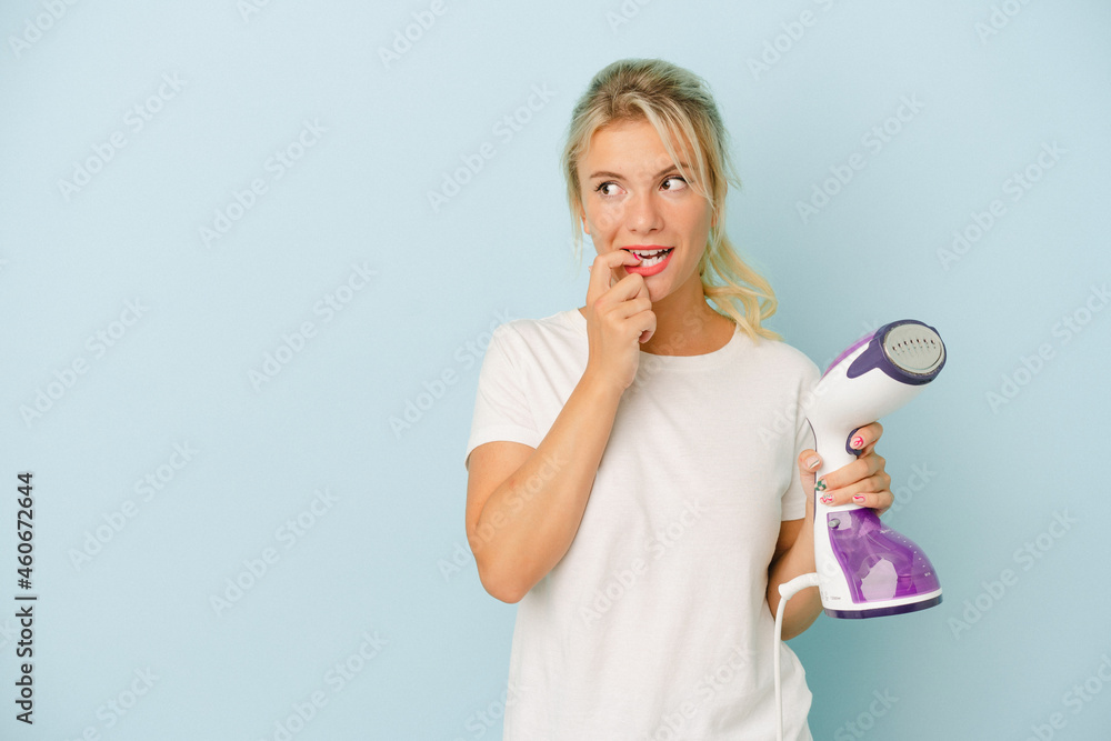 Young Russian woman holding vertical iron isolated on blue background relaxed thinking about something looking at a copy space. Young Russian woman holding vertical iron isolated on blue background relaxed thinking about something looking at a copy space.