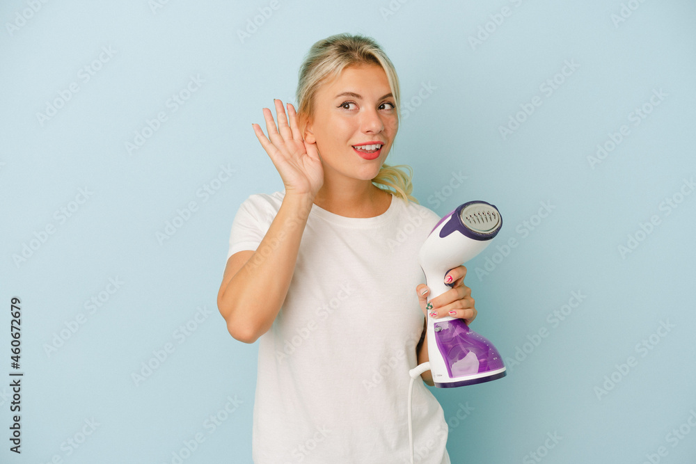 Young Russian woman holding vertical iron isolated on blue background trying to listening a gossip. Young Russian woman holding vertical iron isolated on blue background trying to listening a gossip.