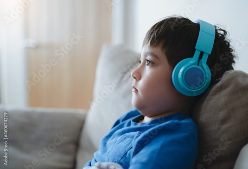 Side view portrait Child boy wearing headphones and looking out deep in thought, School boy in blue T-shirt listening to music,Cute Kid sitting on sofa relaxing in living room at home