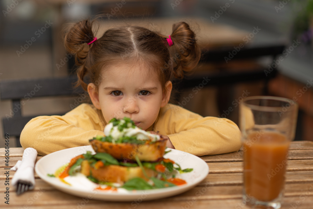 Portrait of little cute angry girl sitting at table in restaurant, against breakfast.Child kid have France lunch outdoors. Concept of eco healthy food.