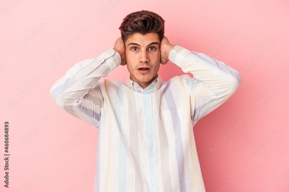 Young caucasian man isolated on pink background covering ears with hands trying not to hear too loud sound. Young caucasian man isolated on pink background covering ears with hands trying not to hear too loud sound.