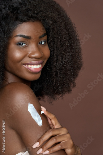 Beautiful smiling black young woman with afro hair and perfect smooth skin looks at camera applying body lotion cream after shower. Skincare, body care and spa concept. Close up vertical portrait.