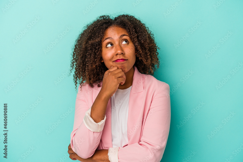 Young african american woman with curly hair isolated on blue background relaxed thinking about something looking at a copy space. Young african american woman with curly hair isolated on blue background relaxed thinking about something looking at a copy space.