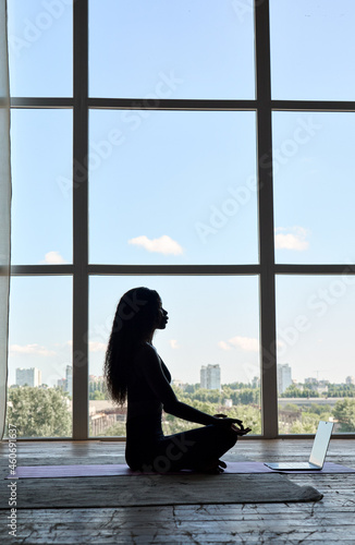 Young peaceful healthy African American black girl silhouette sitting on mat in lotus pose doing yoga exercise remotely watching online class tutorial on laptop pc at home near panoramic window.
