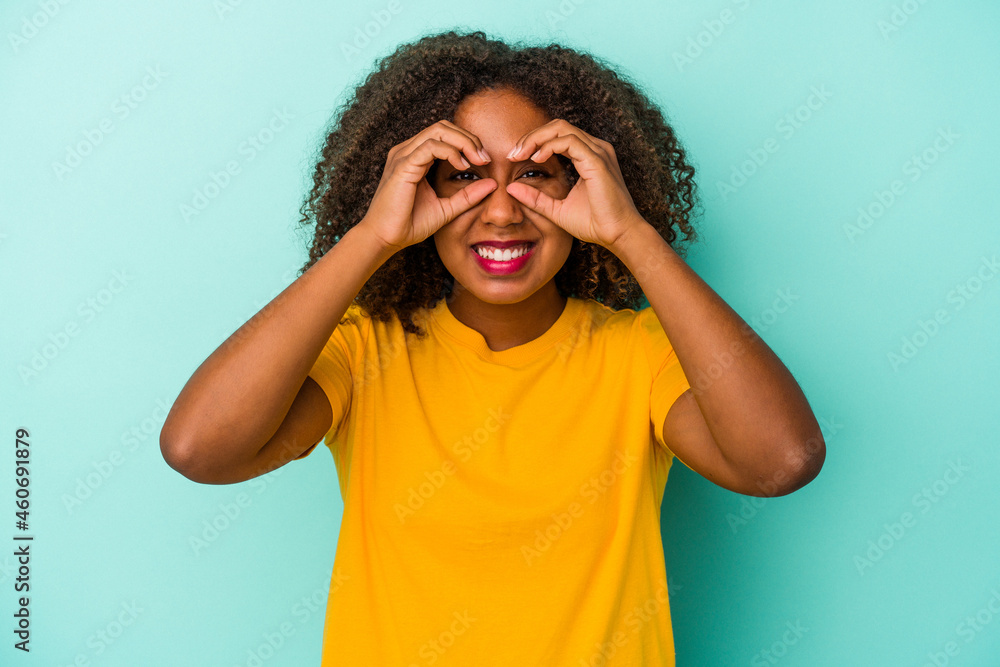 Young african american woman with curly hair isolated on blue background showing okay sign over eyes Young african american woman with curly hair isolated on blue background showing okay sign over eyes
