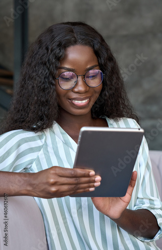 Young happy African black businesswoman student girl in glasses sitting on sofa holding using tablet device surfing dating applications, online chatting, checking work emails at home. Vertical.