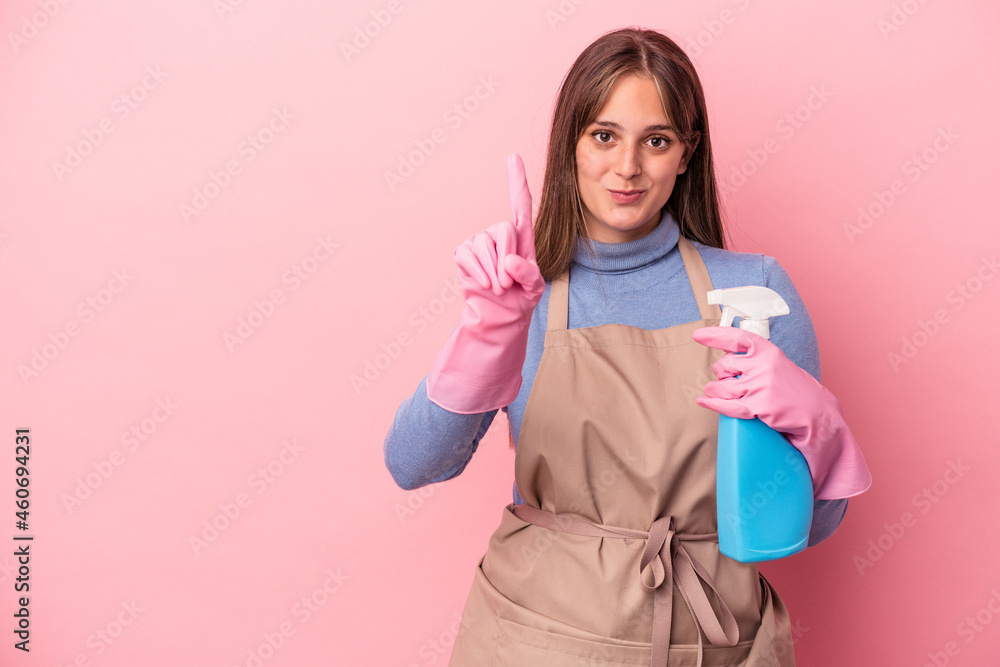 Young caucasian cleaner woman holding spray isolated on pink background showing number one with finger. Young caucasian cleaner woman holding spray isolated on pink background showing number one with finger.