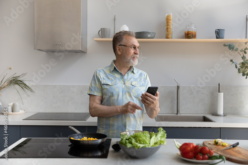 Smiling dreamy middle aged old retired man in eyewear using cellphone  chatting in social network while preparing food for healthy dinner at home  web surfing recipe for meal in modern kitchen.