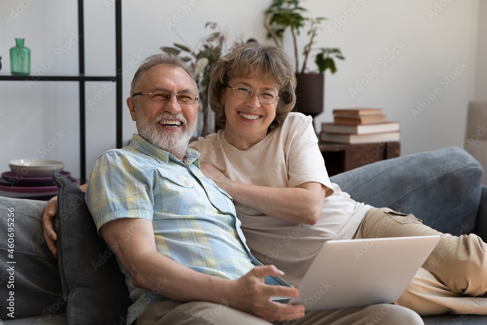 Portrait of smiling beautiful middle aged senior family couple on eyeglasses relaxing on comfortable sofa with computer on laps, enjoying leisure weekend time together at home, older people pastime.