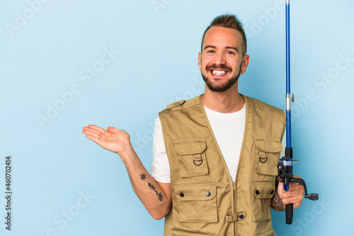 Young caucasian fisherman with tattoos holding rod isolated on blue background showing a copy space on a palm and holding another hand on waist.