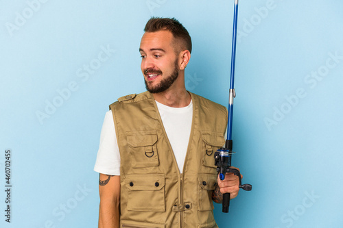 Young caucasian fisherman with tattoos holding rod isolated on blue background looks aside smiling, cheerful and pleasant.