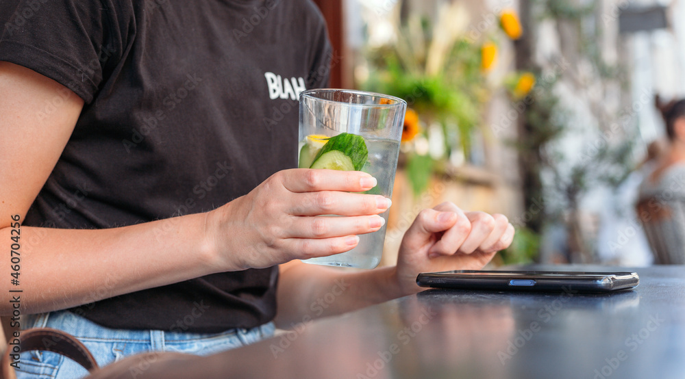 Woman at cafe drinking water and using mobile phone. Hand holding lemonade on cafe background. Glass of Cold drinks. Street front view, girl waiting for her order Woman at cafe drinking water and using mobile phone. Hand holding lemonade on cafe background. Glass of Cold drinks. Street front view, girl waiting for her order