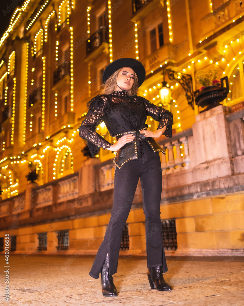 Christmas lights in the city, blonde model dressed in black with a hat next to an illuminated building in a fashionable pose, winter lifestyle