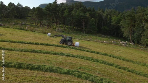 Tractor With Bale Graber Collecting Hay Bale Wrapped In White Plastic Film On Farm. wide photo