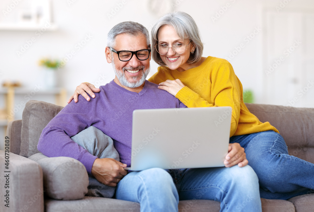 Beautiful mature family couple looking at laptop with smile on face while spending time together Beautiful mature family couple looking at laptop with smile on face while spending time together