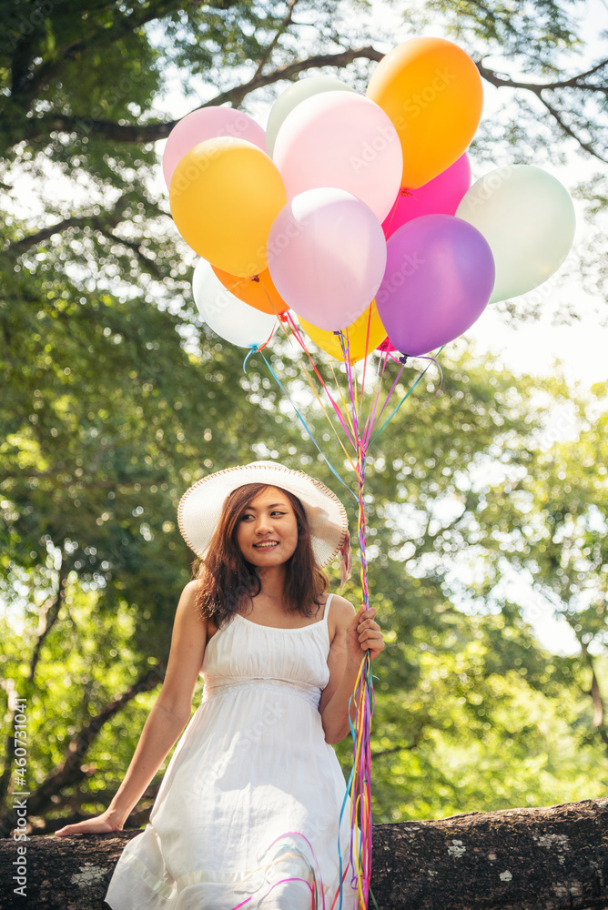 Cheerful beauty woman holding balloons relax sitting under big tree in green park with happiness. Woman Hands holding vibrant air balloons play on birthday party happy time summer on sunshine outdoor