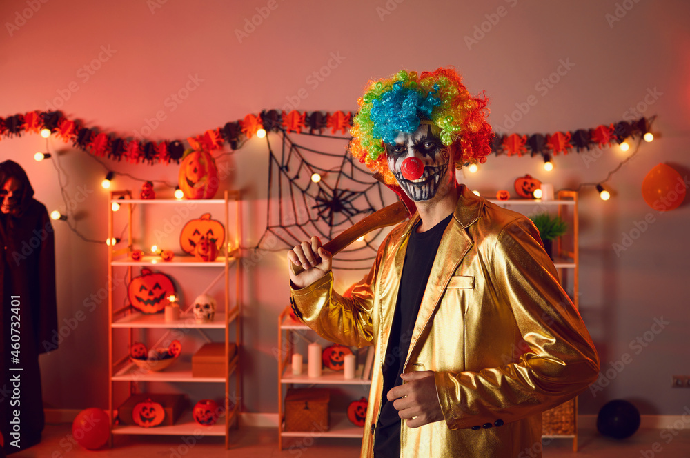 Indoor portrait of guy in costume of spooky evil clown at Halloween party. Adult man wearing gold suit, colorful wig and fake red nose, with creepy makeup on face holding axe and looking at camera