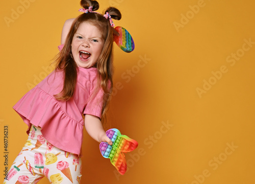 Screaming redhead kid girl in pink shirt and colorful pants with lollipop girlish print dances holding two sensory rainbow color toys, round and butterfly pop in hands over background with copy space