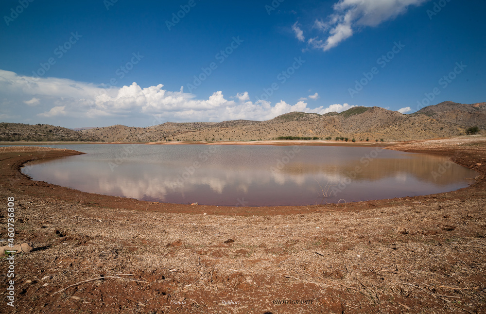 Oval shaped lake in Gandiali Dam in Pakistan with reflection of clouds in the sky, mountains in the background and red soil in foreground