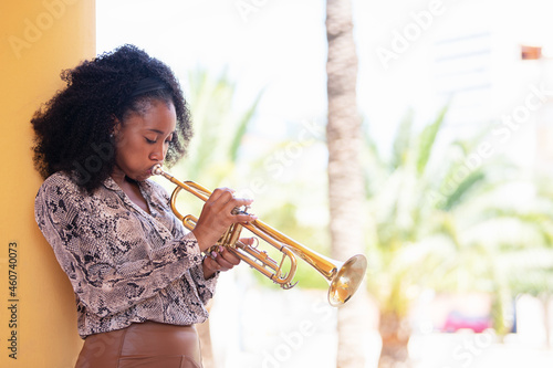 Concentrated afro american woman leaning against a concrete column playing the trumpet outside