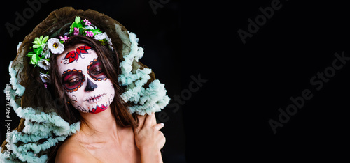 Fototapeta Naklejka Na Ścianę i Meble -  A young girl with dark hair and sugar skull makeup with flowers on her head looks into the camera on a black background. Panoramic stretched image for banner