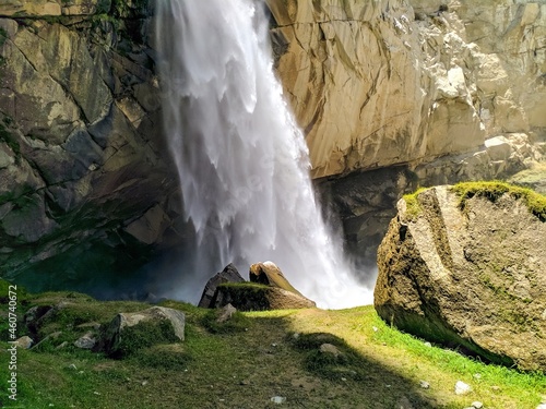Waterfall falling from above the mountains with speed falling in the stream along with rocks in surrounding and a large stone with grass grown over it in the foreground.