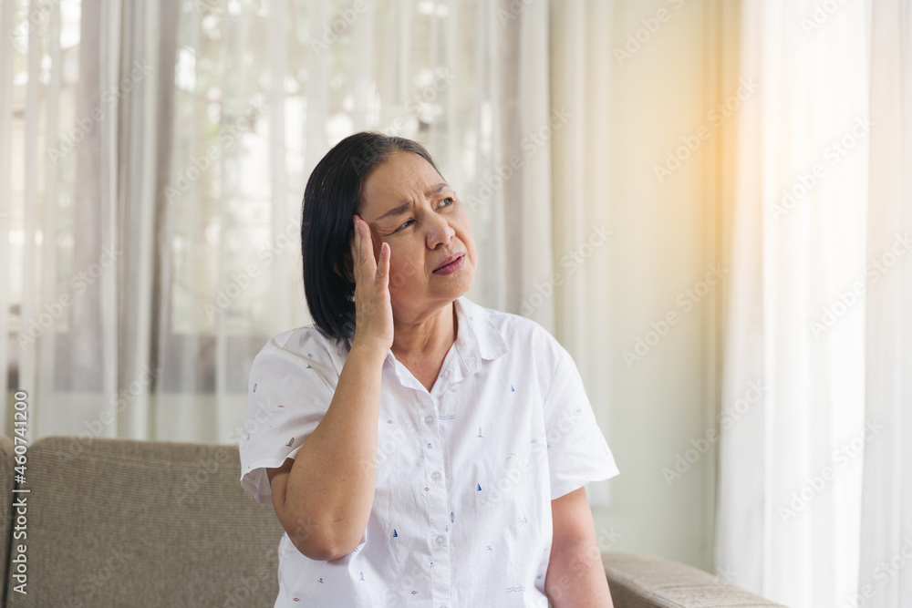 Elderly Asian woman sitting and having a headache and touching her head with her hands at home. Retirement, quarantine and health care concept Elderly Asian woman sitting and having a headache and touching her head with her hands at home. Retirement, quarantine and health care concept