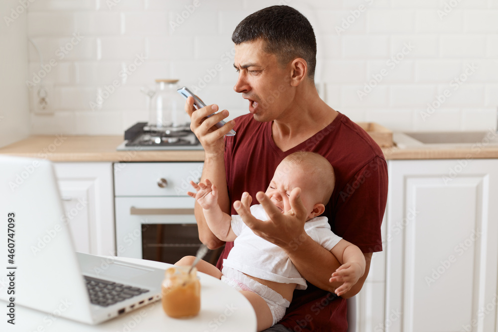 Angry aggressive brunette male wearing maroon casual style t shirt, sending voice message, screaming on telephone, sitting at table in kitchen with his infant daughter. Angry aggressive brunette male wearing maroon casual style t shirt, sending voice message, screaming on telephone, sitting at table in kitchen with his infant daughter.