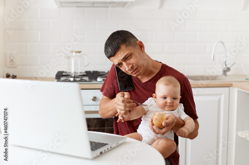 Stressed sad brunette male wearing maroon casual style t shirt sitting at table in kitchen, having conversation via smart phone, having sorrow look, feeding his infant daughter with fruit puree. photo