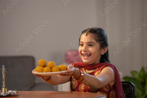 A small girl dressed In traditional cloth happily offering a plate of ladoos. photo