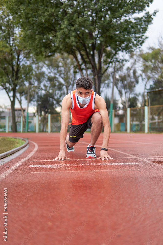 Young latin guy with protective mask, in position to run at full speed on a track, ready, at the start line, copy space