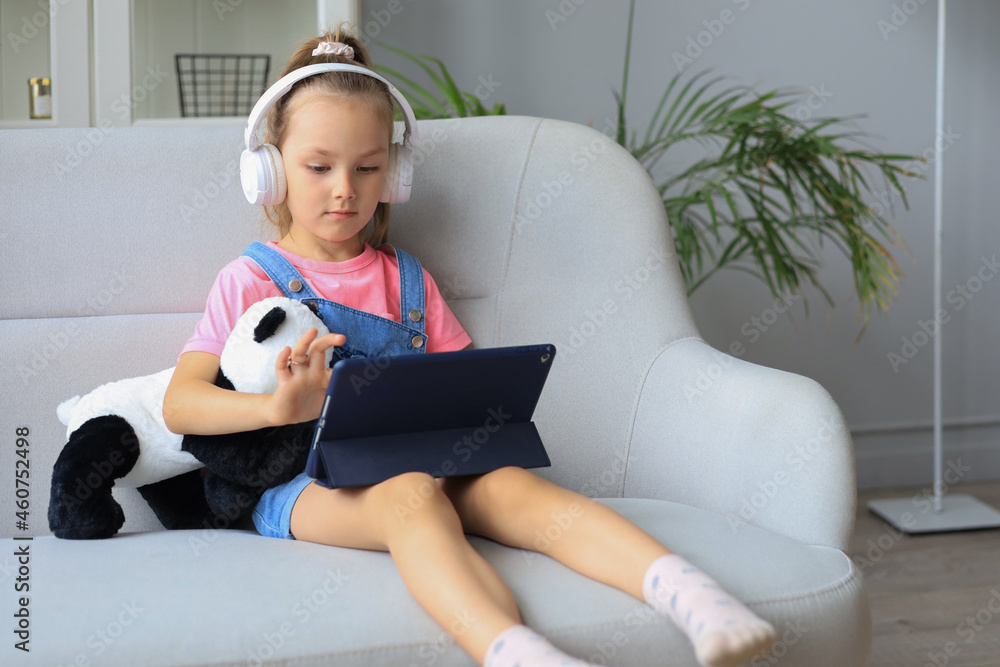 Smiling little girl sitting on sofa playing online games, web surfing information, using funny applications on tablet. Smiling little girl sitting on sofa playing online games, web surfing information, using funny applications on tablet.
