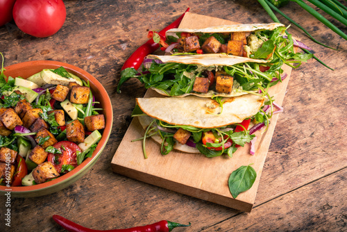 Tacos with vegetable salad and fried tofu on a wooden board on a table with a bowl of salad nearby photo