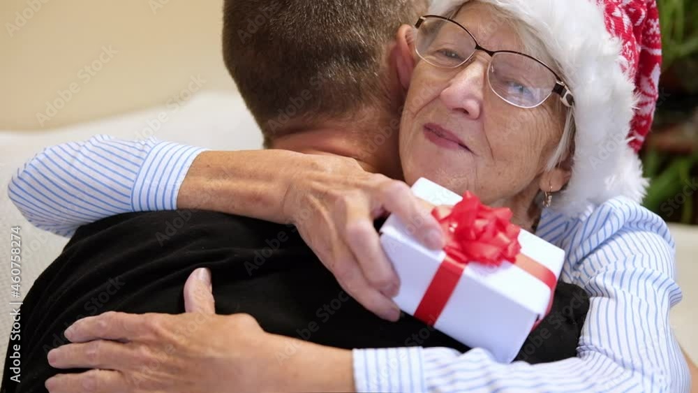 Young man embracing his senior mother and giving Christmas present. Mother hugging son Christmas eve. Elderly woman having gift surprise from son. family celebrating new year together on evening home Young man embracing his senior mother and giving Christmas present. Mother hugging son Christmas eve. Elderly woman having gift surprise from son. family celebrating new year together on evening home