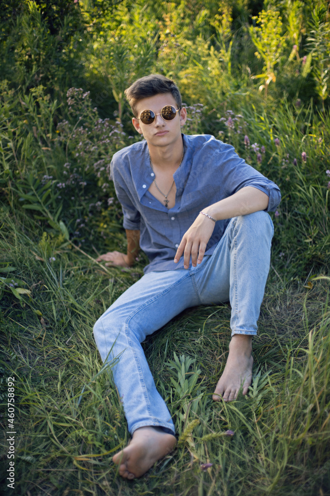 sexy teen guy in sunglasses is sitting on the grass in nature in a shirt and jeans sexy teen guy in sunglasses is sitting on the grass in nature in a shirt and jeans