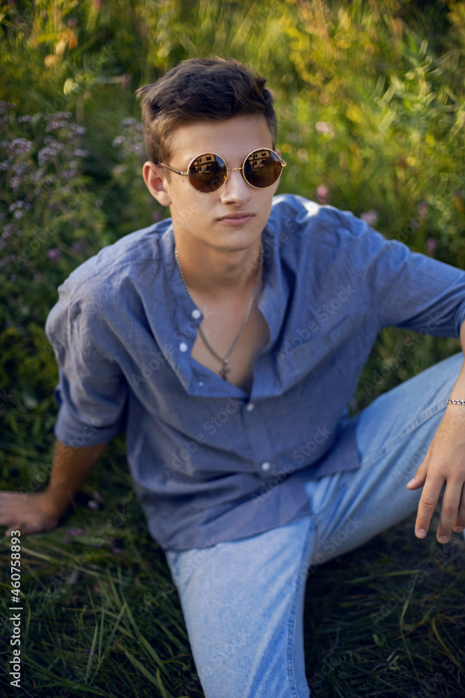 sexy teen guy in sunglasses is sitting on the grass in nature in a shirt and jeans sexy teen guy in sunglasses is sitting on the grass in nature in a shirt and jeans