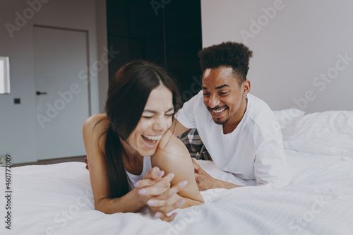 Laughing young couple two family in casual white clothes lying in bed on stomach man tickling woman relax spend time together in bedroom lounge home house wake up dream be lost in reverie good day photo