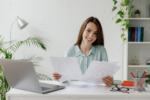 Smiling happy young successful employee business woman in blue shirt hold paper account documents sit work at workplace white desk with laptop pc computer at office indoors Achievement career concept photo