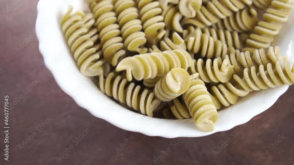 Full white bowl of fusilli Italian uncooked pasta on a wooden surface. Falling green pea vegetable spiral macaroni in slow motion. Macro. Mediterranean cuisine concept. Rotation Full white bowl of fusilli Italian uncooked pasta on a wooden surface. Falling green pea vegetable spiral macaroni in slow motion. Macro. Mediterranean cuisine concept. Rotation