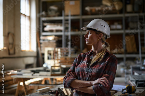 Portrait of female engineer looking aside with arms crossed indoors in carpentry workshop. Small business concept.