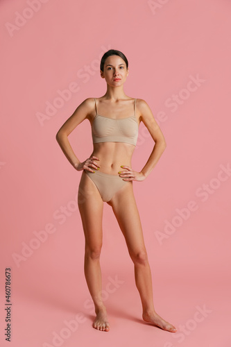 Portrait of young attractivegirl, slim woman in nude color lingerie, bra and panties posing isolated over pink studio background. © master1305
