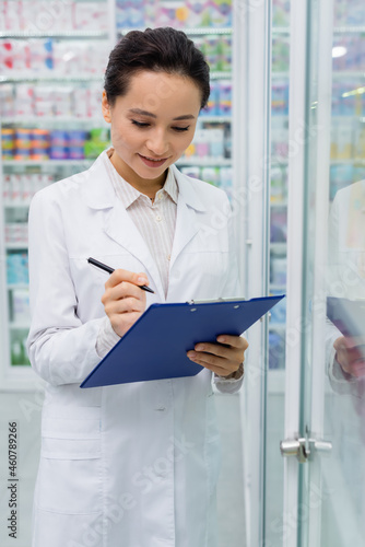 cheerful pharmacist in white coat writing on clipboard © LIGHTFIELD STUDIOS