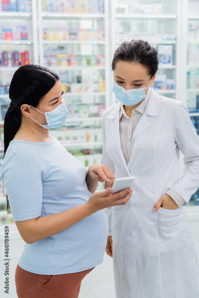 asian pregnant woman in medical mask pointing at smartphone near pharmacist in drugstore