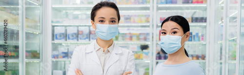 pharmacist in medical mask standing near asian customer in drugstore, banner