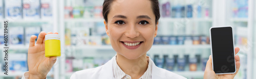 cheerful pharmacist in white coat holding smartphone with blank screen and bottle in drugstore