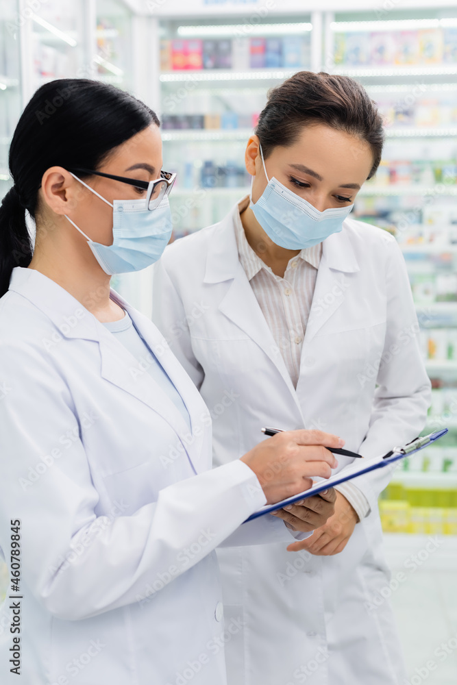 asian pharmacist in glasses and medical mask writing on clipboard near colleague asian pharmacist in glasses and medical mask writing on clipboard near colleague
