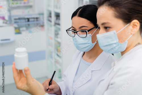 interracial pharmacists in medical masks looking at bottle in drugstore